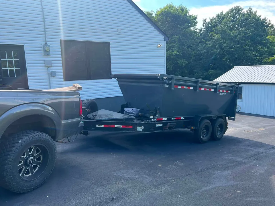 TX2 truck backing a gray roll-off dumpster onto a driveway beside a white house